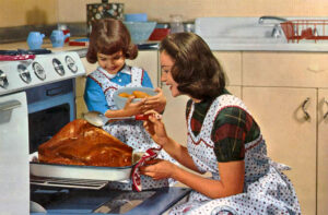smiling woman with pinafore apron pulls turkey from oven as little girl in apron looks on